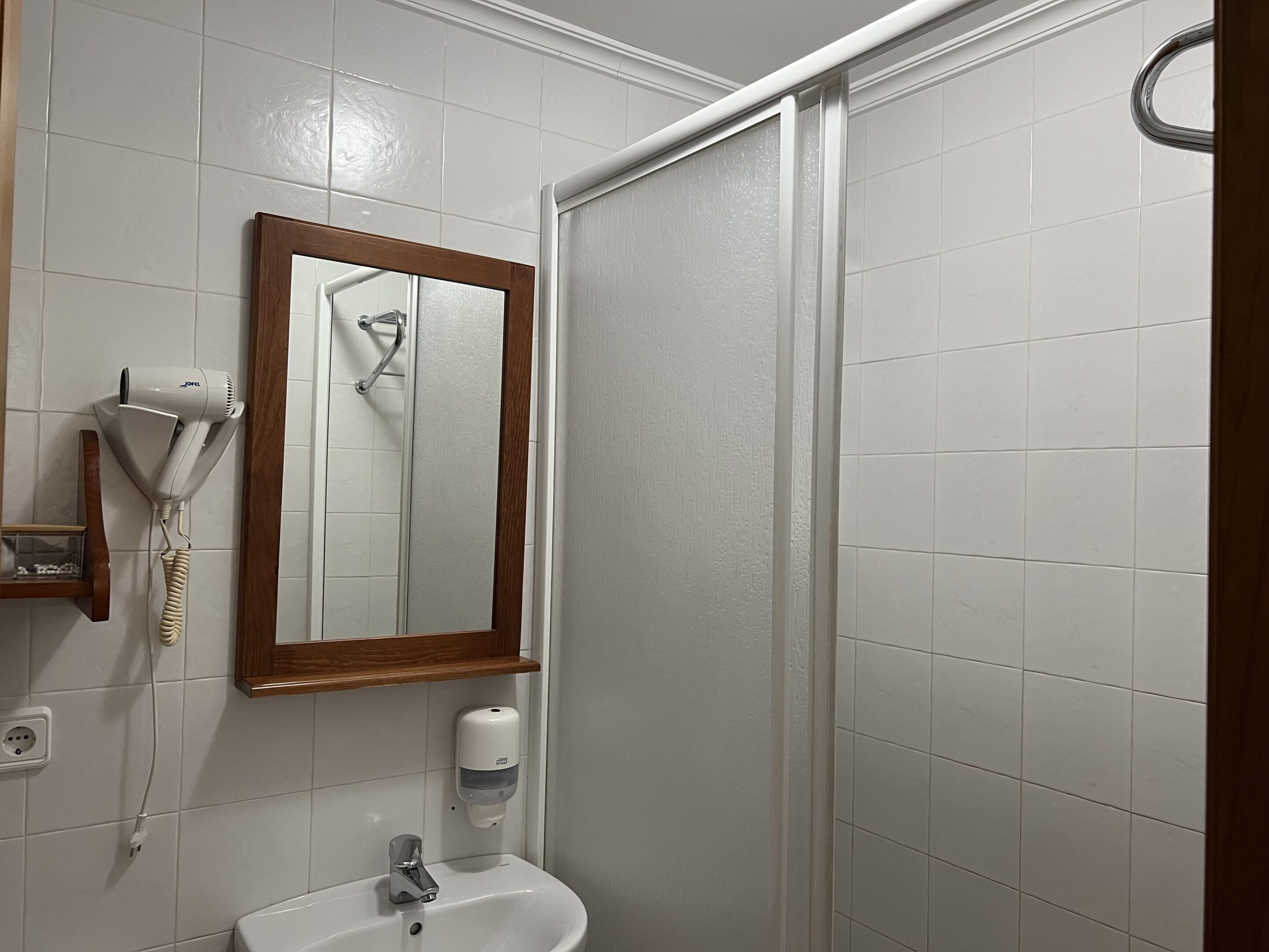 Une salle de bain avec un lavabo blanc, un sèche-cheveux, un miroir et un espace douche.