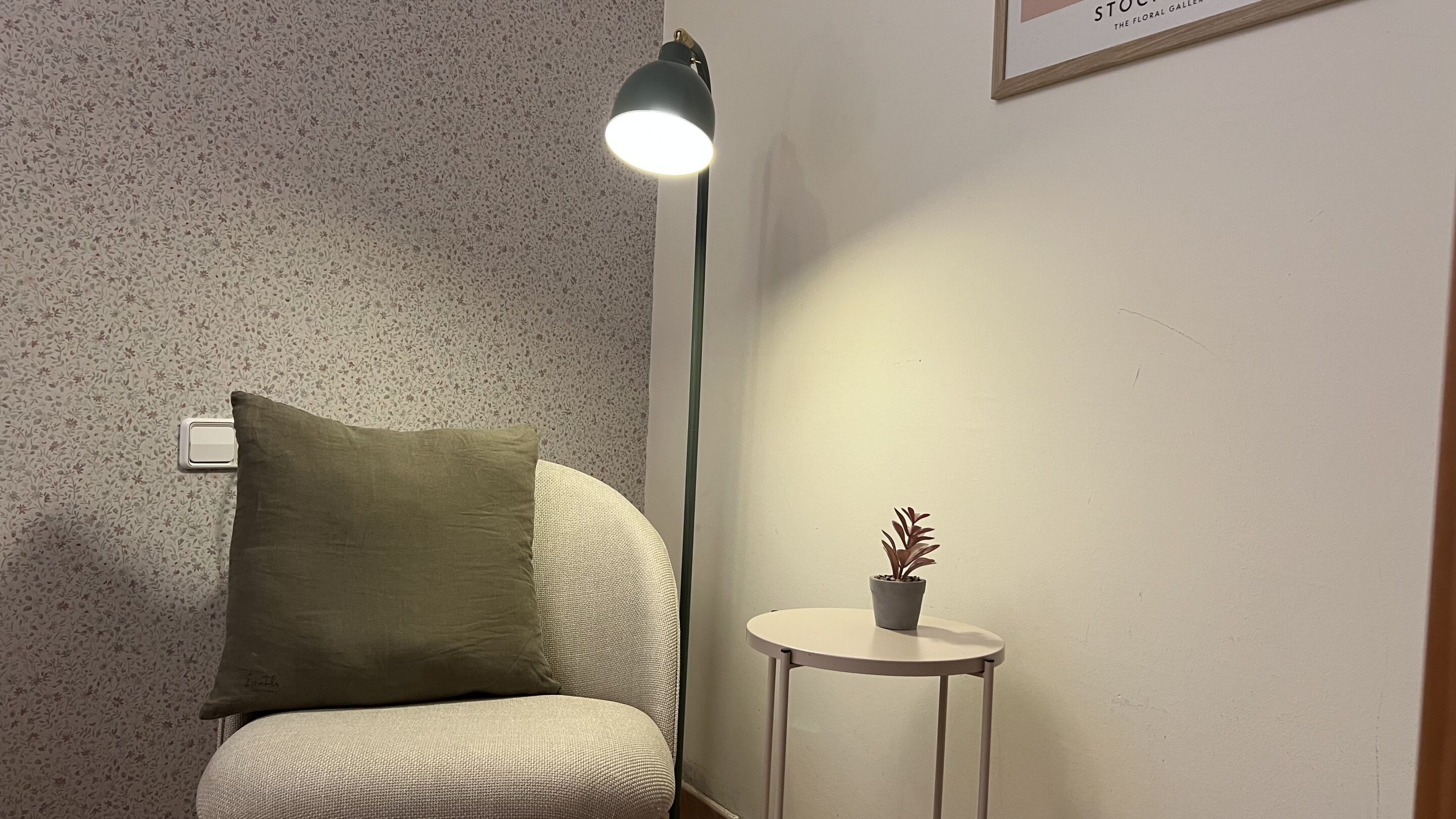 A corner of a room with a patterned wall, a green pillow on a chair, a floor lamp, and a small table with a potted plant.