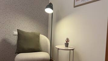 Rincón de una habitación con un papel tapiz estampado, un cojín verde sobre una silla, una lámpara de pie y una mesita con una planta en maceta.