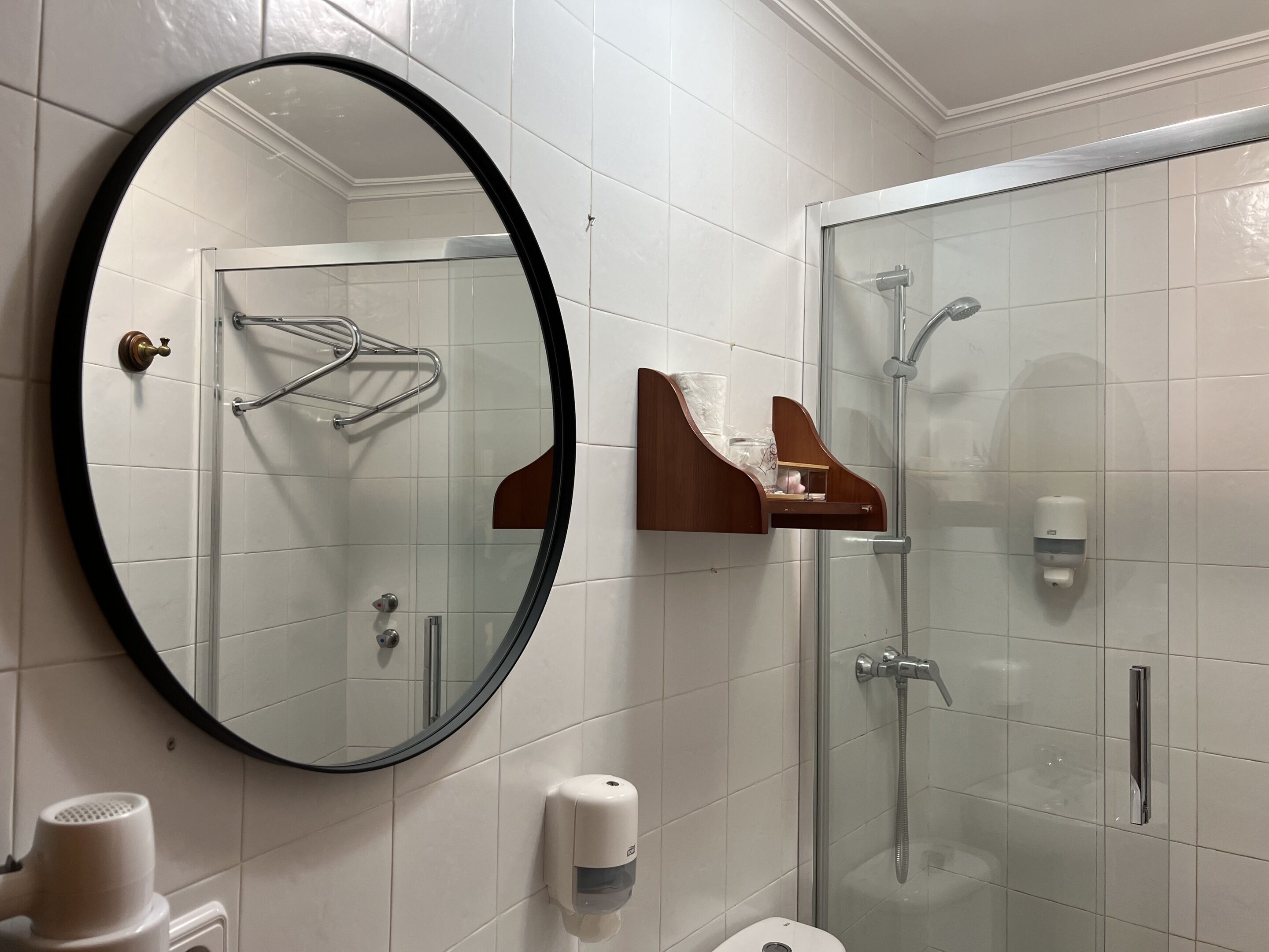 A bathroom with a round mirror, a glass-enclosed shower, a towel rack, and a soap dispenser.