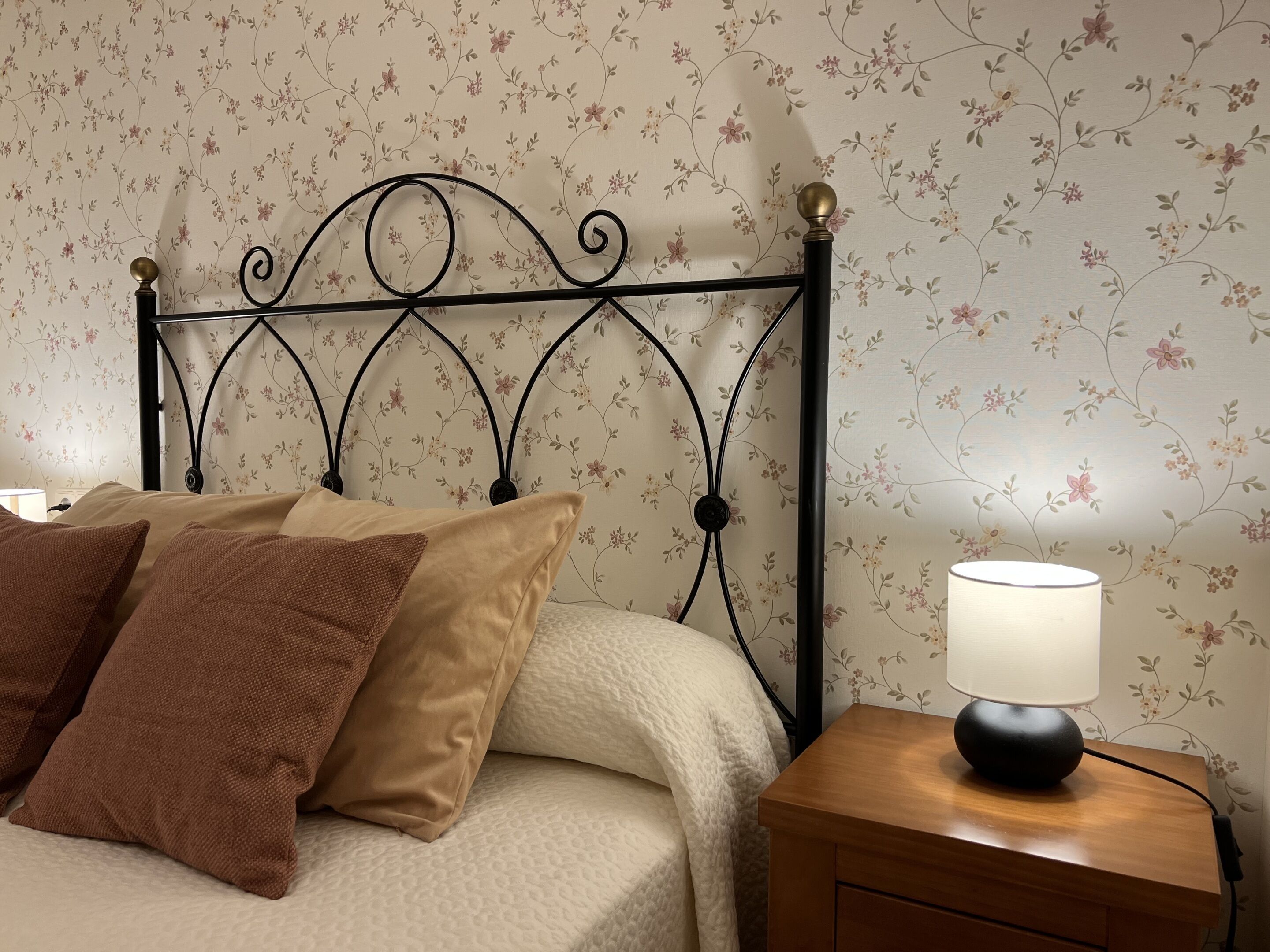 Una cama con papel tapiz floral, un cabecero metálico y una mesita de noche con una lámpara.