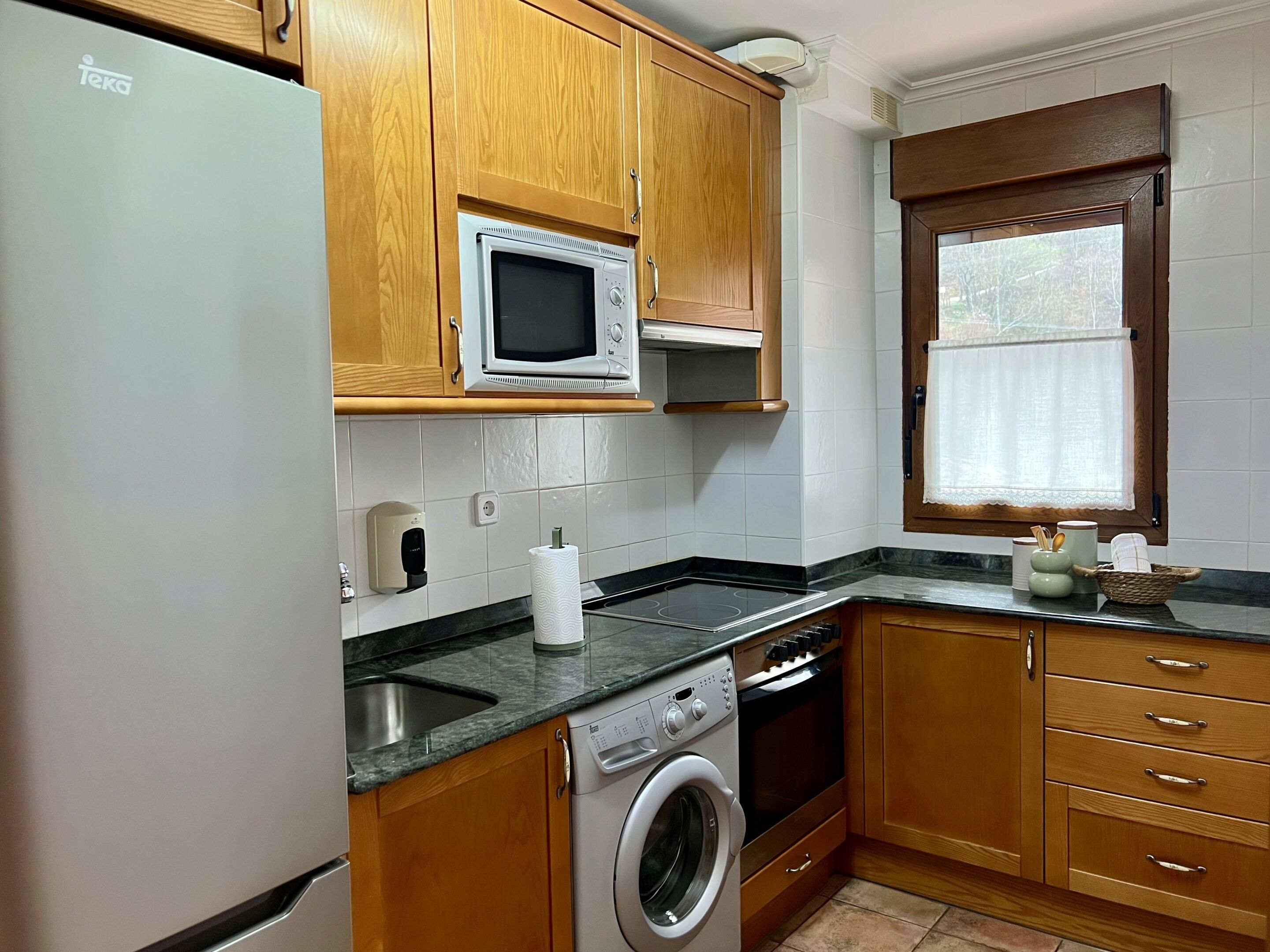 A kitchen with wooden cabinets, a built-in microwave, a washing machine, and a window with a curtain.