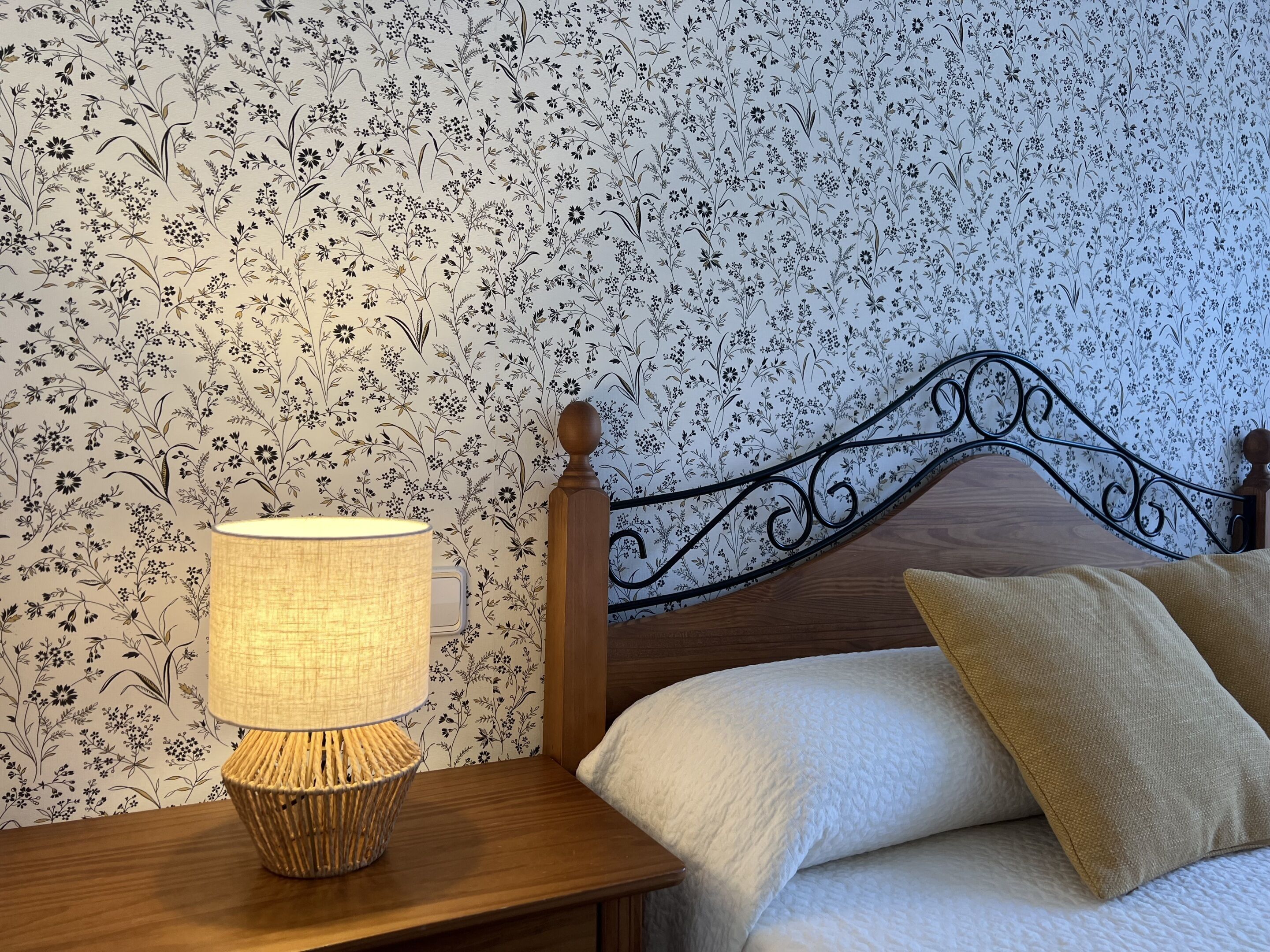 Een netjes opgemaakt bed met een houten hoofdeinde, een bedlamp en behang met een bloemenmotief.