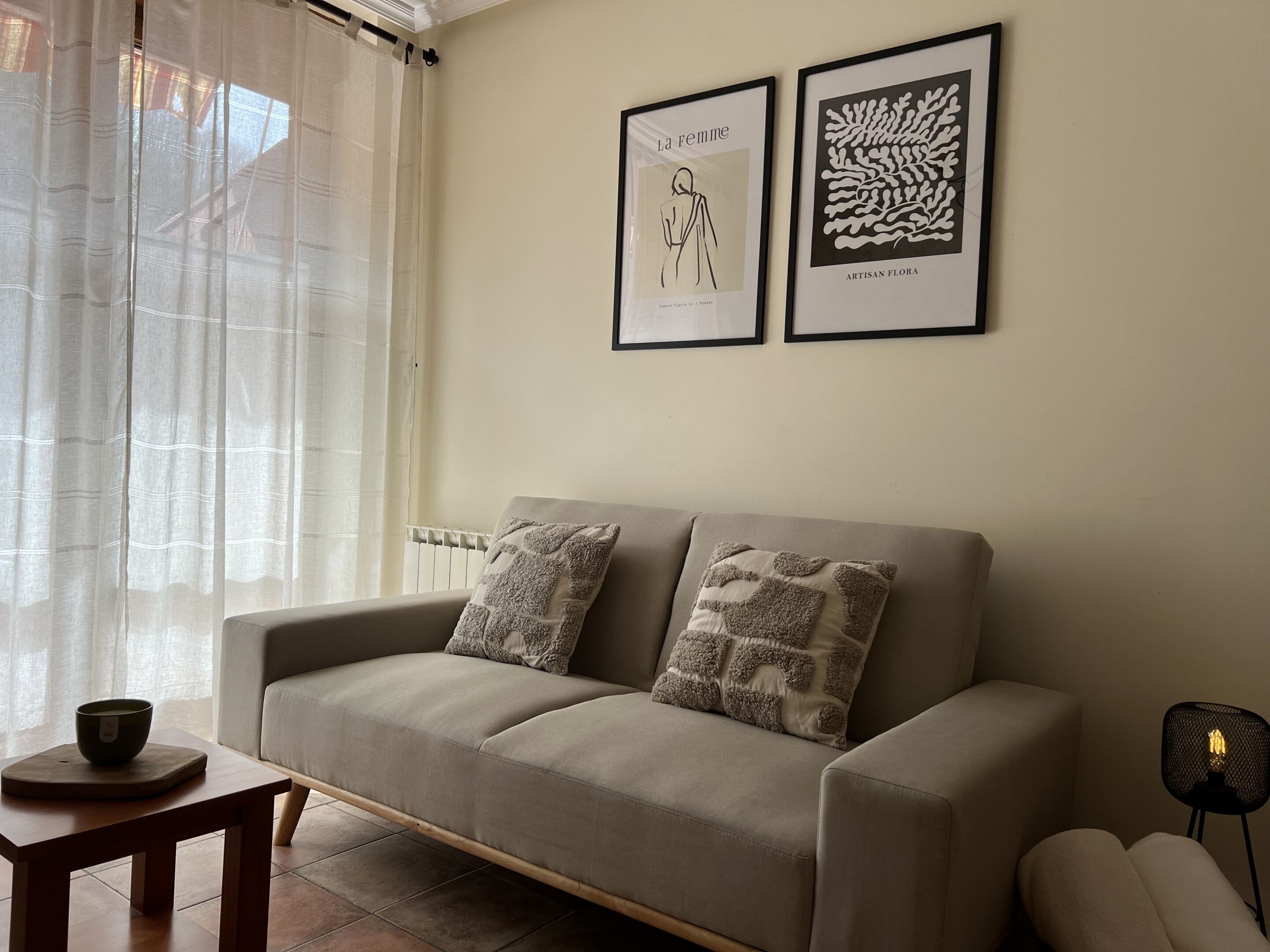 Un salon avec un canapé beige, deux coussins à motifs, une petite table en bois sur laquelle se trouve un bol, et deux œuvres d’art encadrées accrochées au mur.