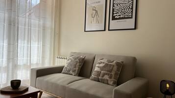 Un salon avec un canapé beige, deux coussins à motifs, une petite table en bois sur laquelle se trouve un bol, et deux œuvres d’art encadrées accrochées au mur.