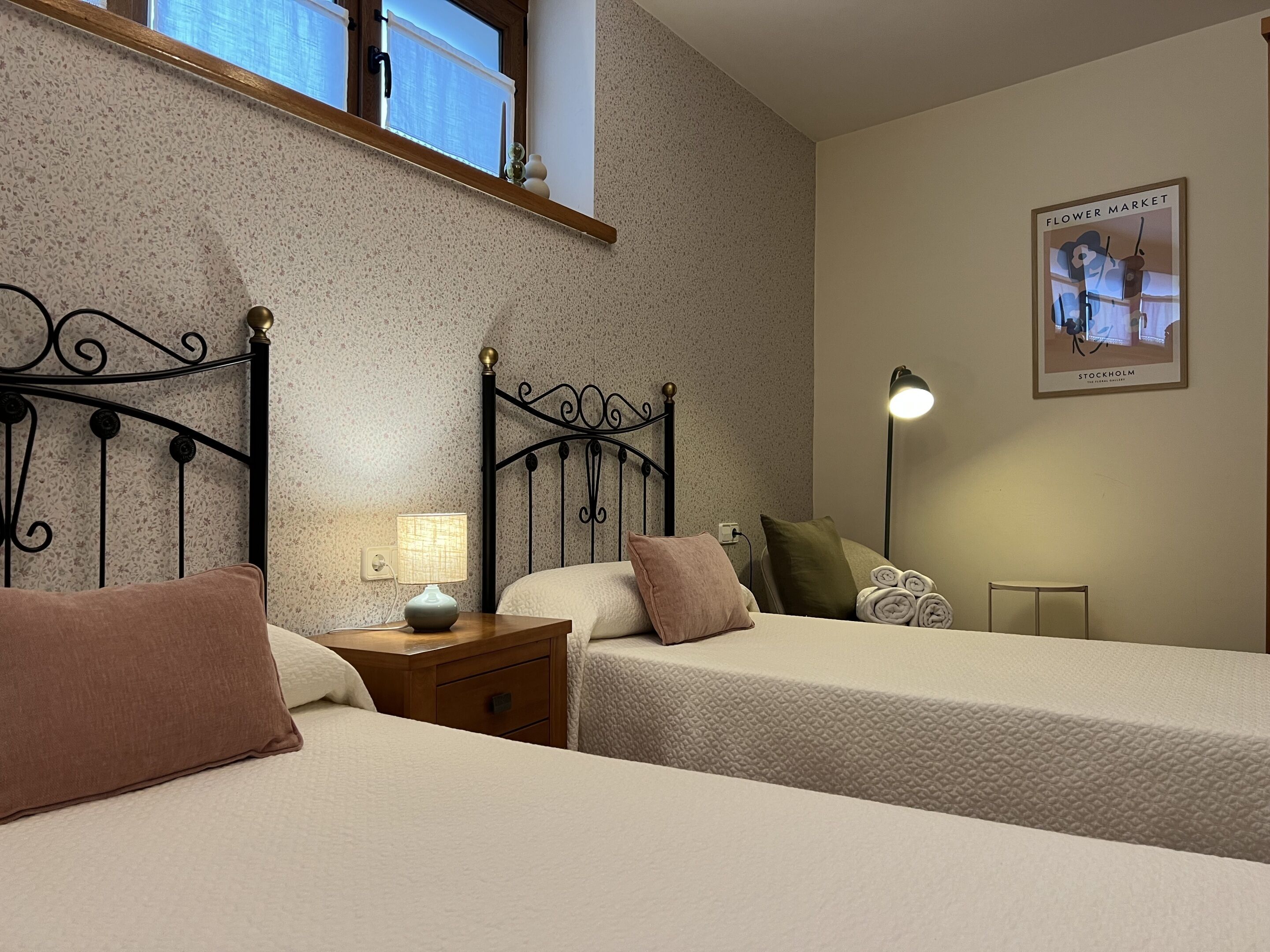 Una habitación de hotel con dos camas, una mesita de noche con lámpara, un cuadro en la pared y una ventana con vistas al cielo.