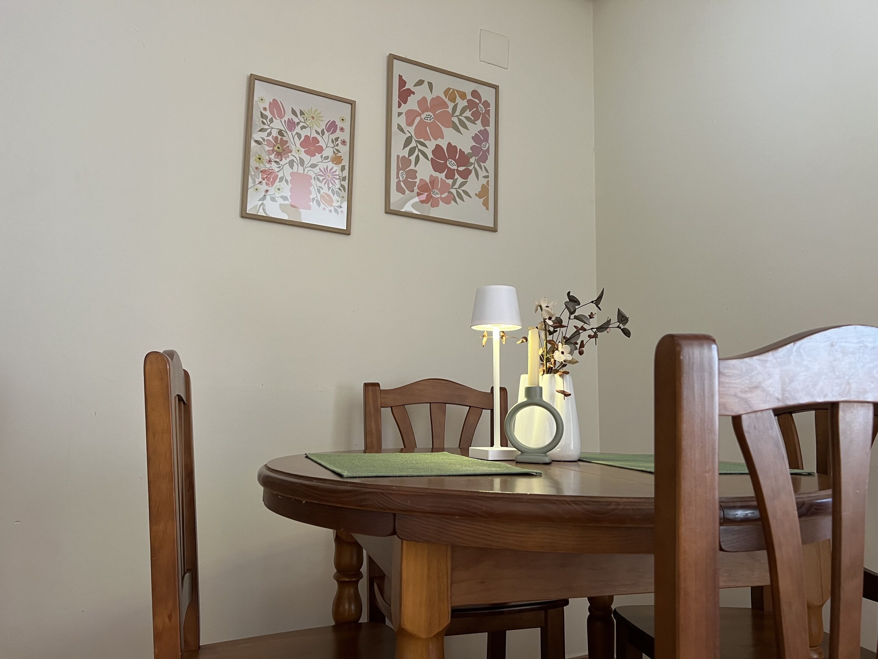 Taula de menjador amb caires, una làmpada i dos quadres florals al mur.