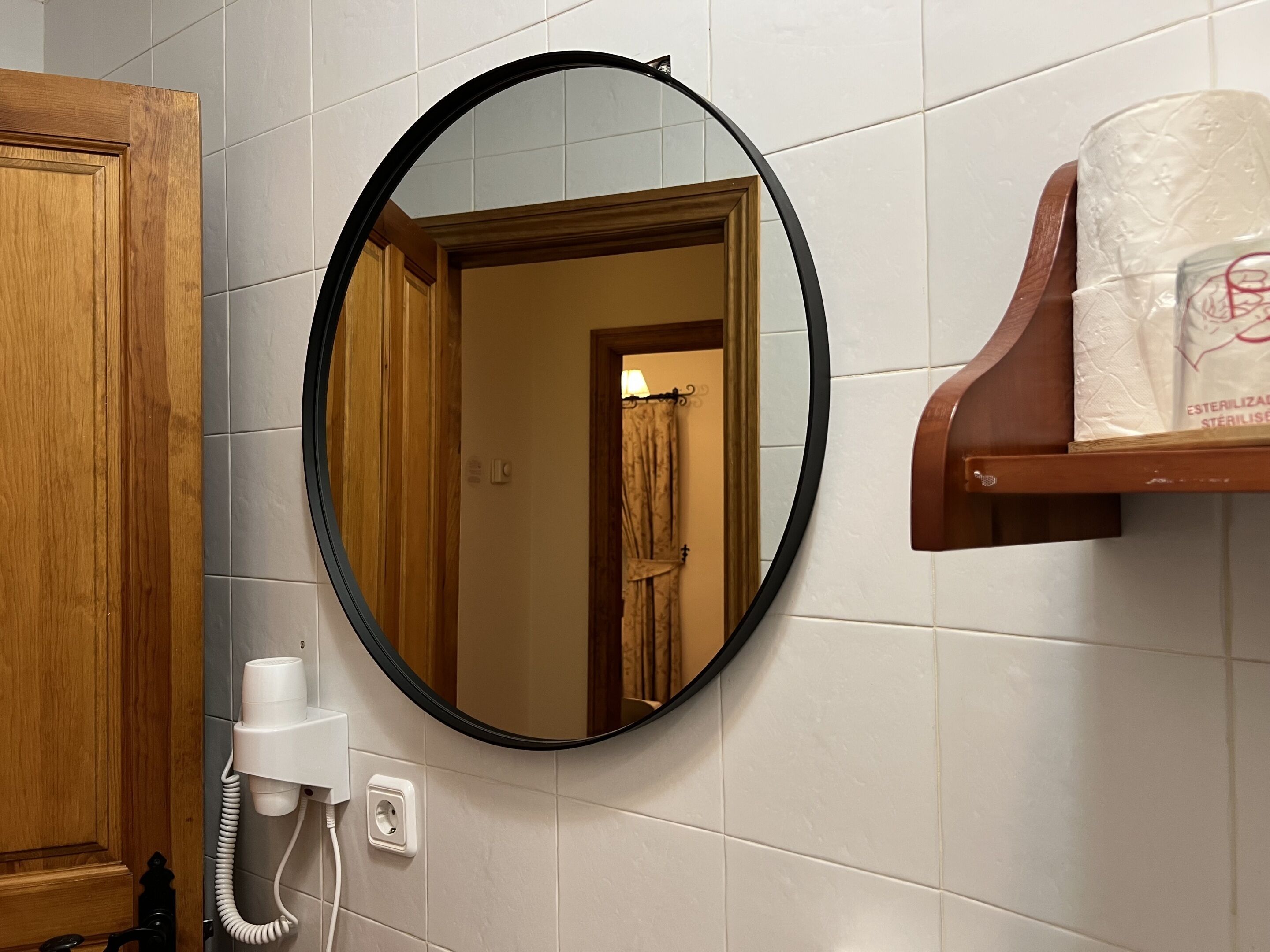 Et badeværelse med en trædør, et rundt spejl, en håndklædeholder og en toiletpapirholder.