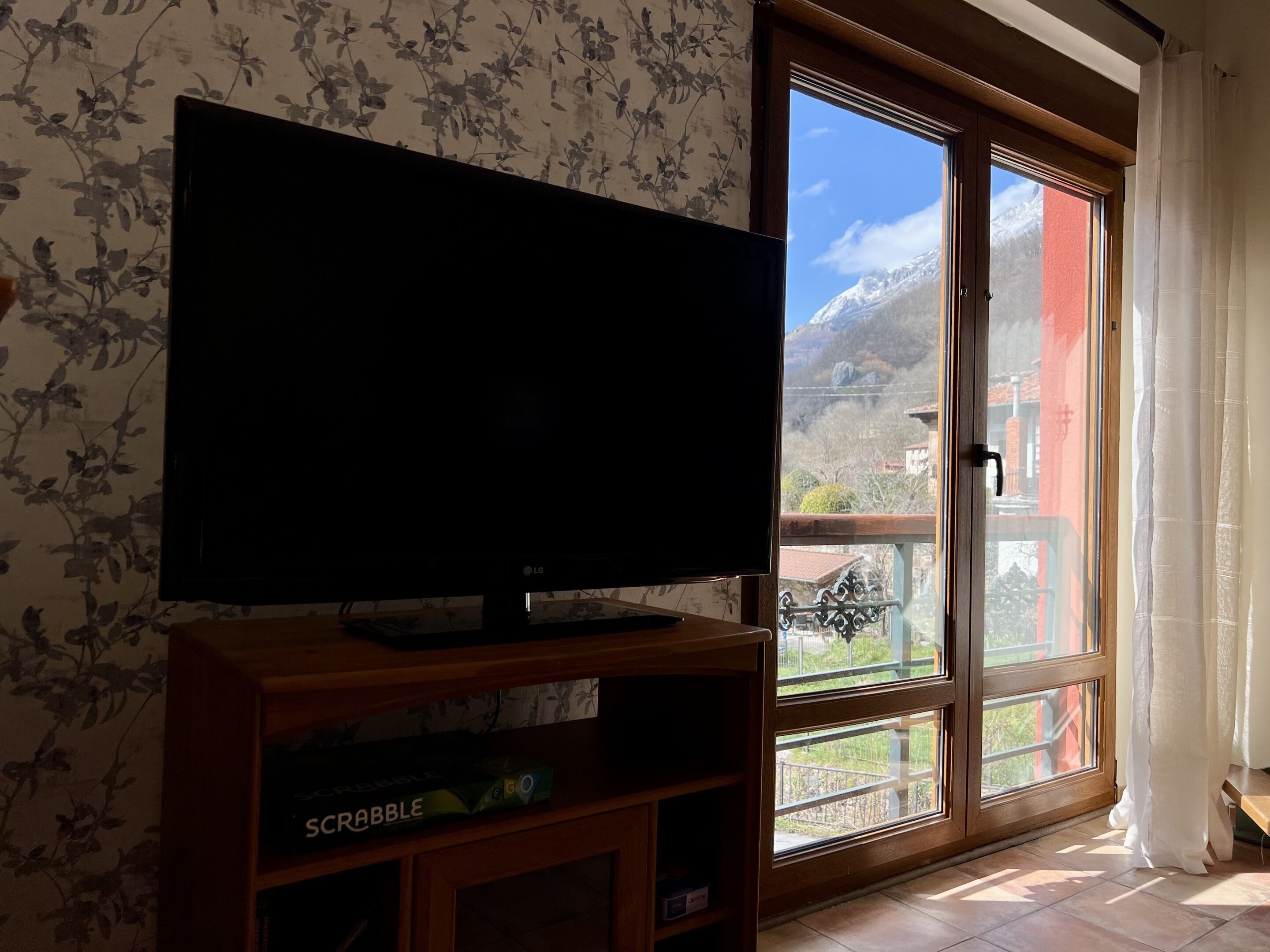 A room with a television on a wooden stand, a window with a view of mountains, and a balcony with a railing.