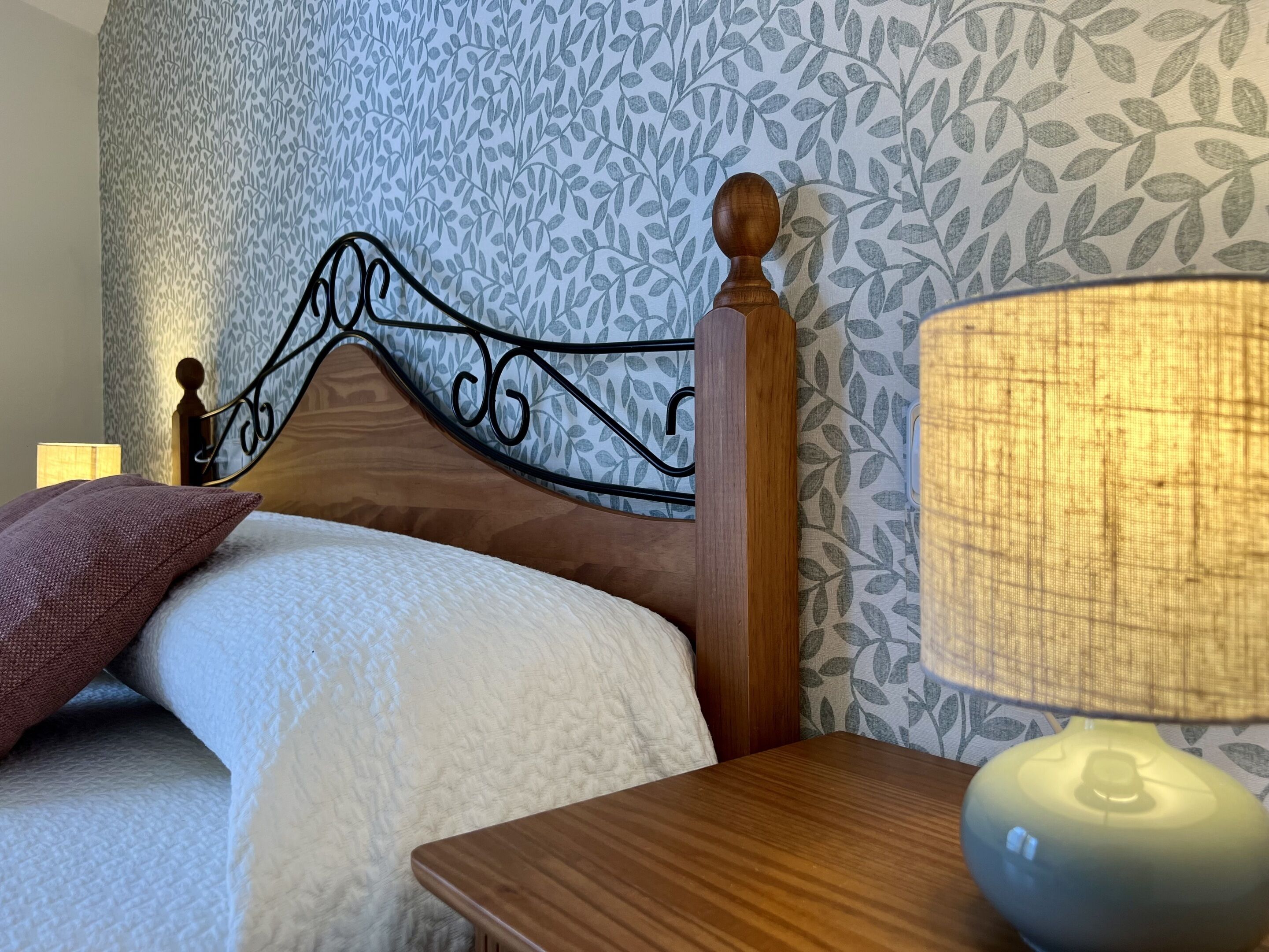 A neatly made bed with a wooden headboard, a bedside lamp, and patterned wallpaper.