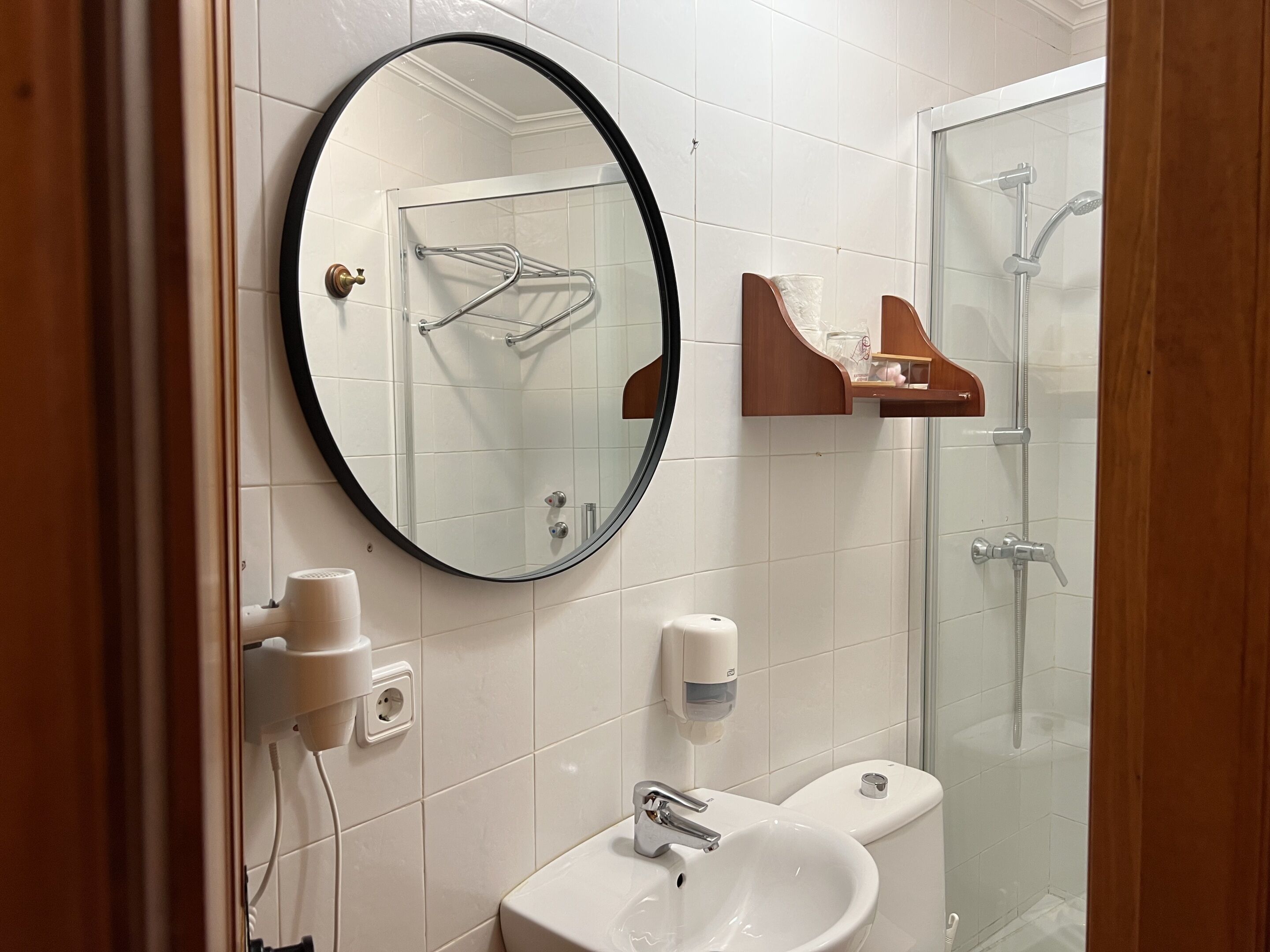 Un baño con un espejo redondo, un lavamanos, un inodoro y una ducha con mampara de vidrio.