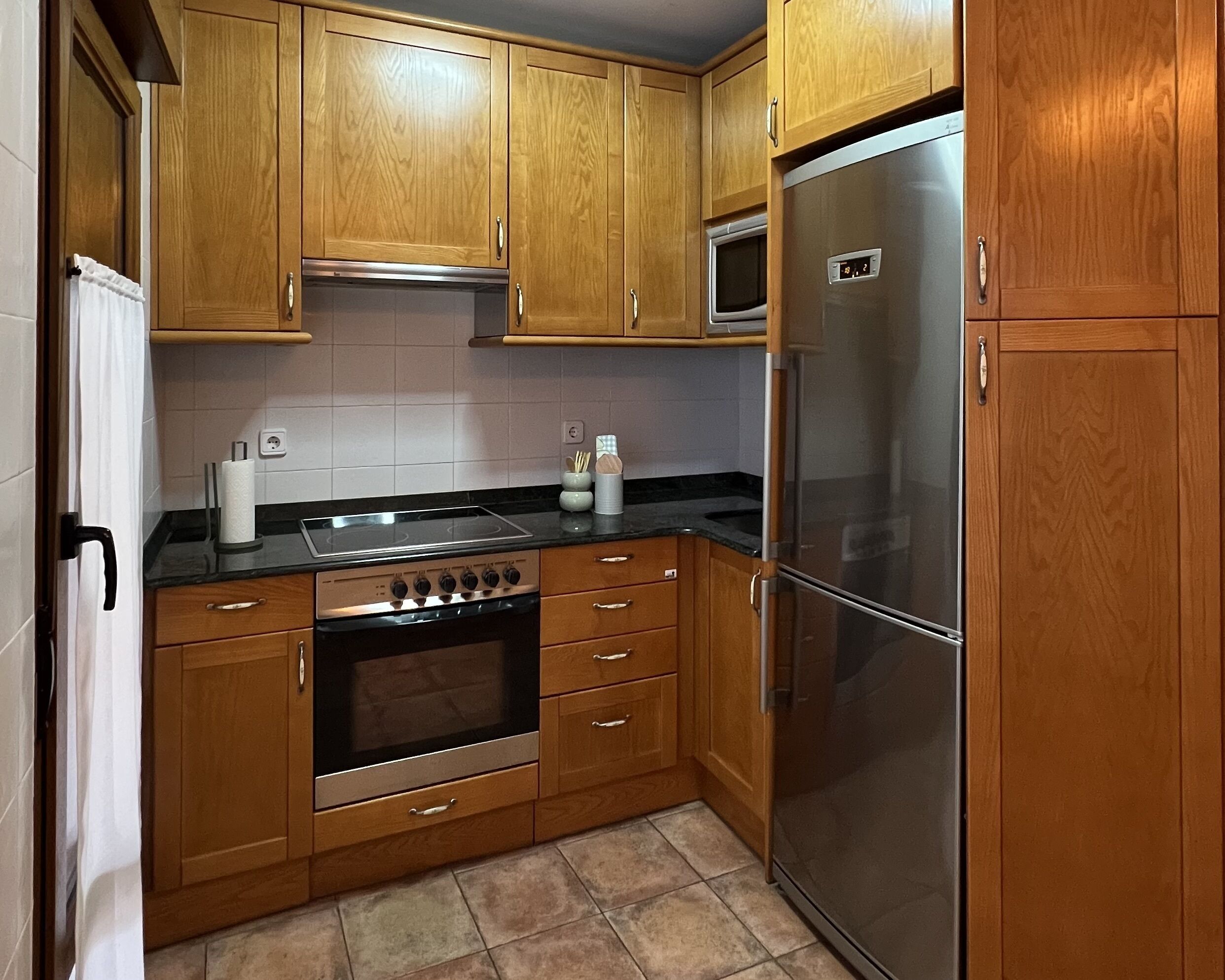 A compact kitchen with wooden cabinets, a stainless steel refrigerator, a built-in oven, and a microwave.