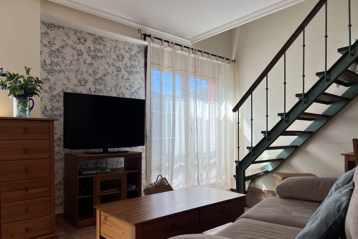 Een woonkamer met een houten tv-meubel, een flatscreen televisie, een houten salontafel, een beige bank en een trap met een houten leuning.