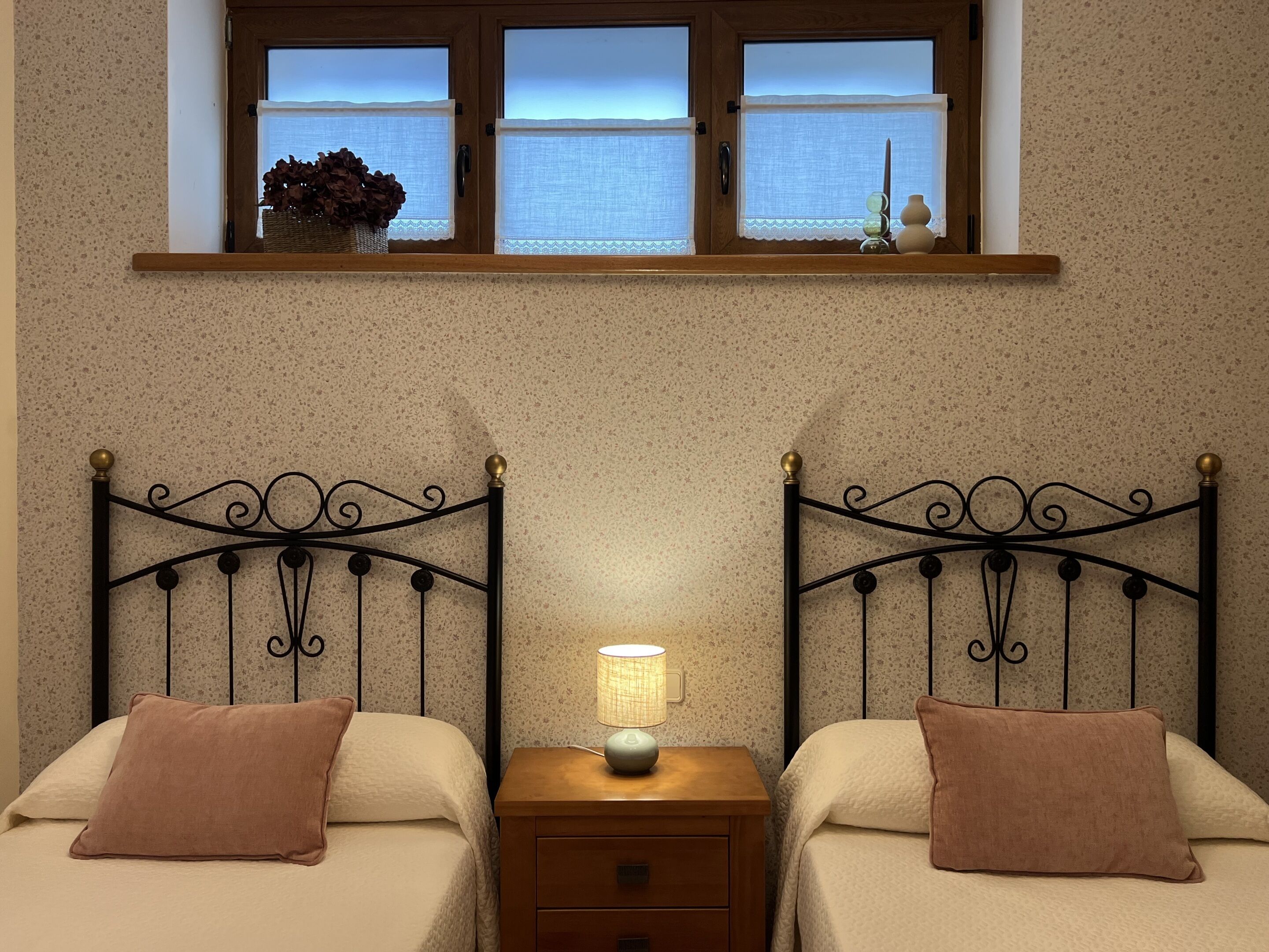 A bedroom with two beds, a nightstand with a lamp, and a window with a view.