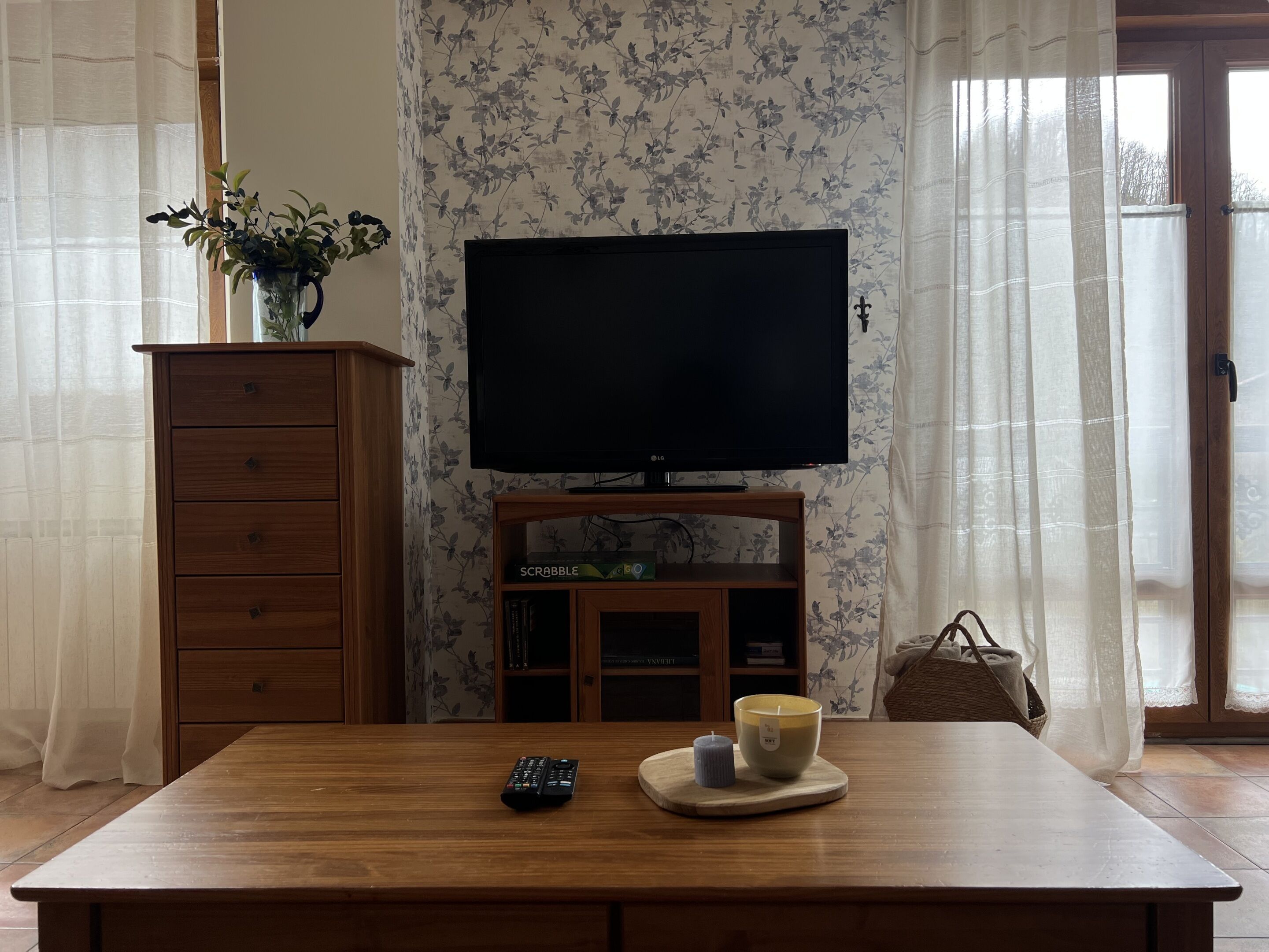 A living room with a wooden table, a television on a stand, a vase with plants, and a bookshelf.