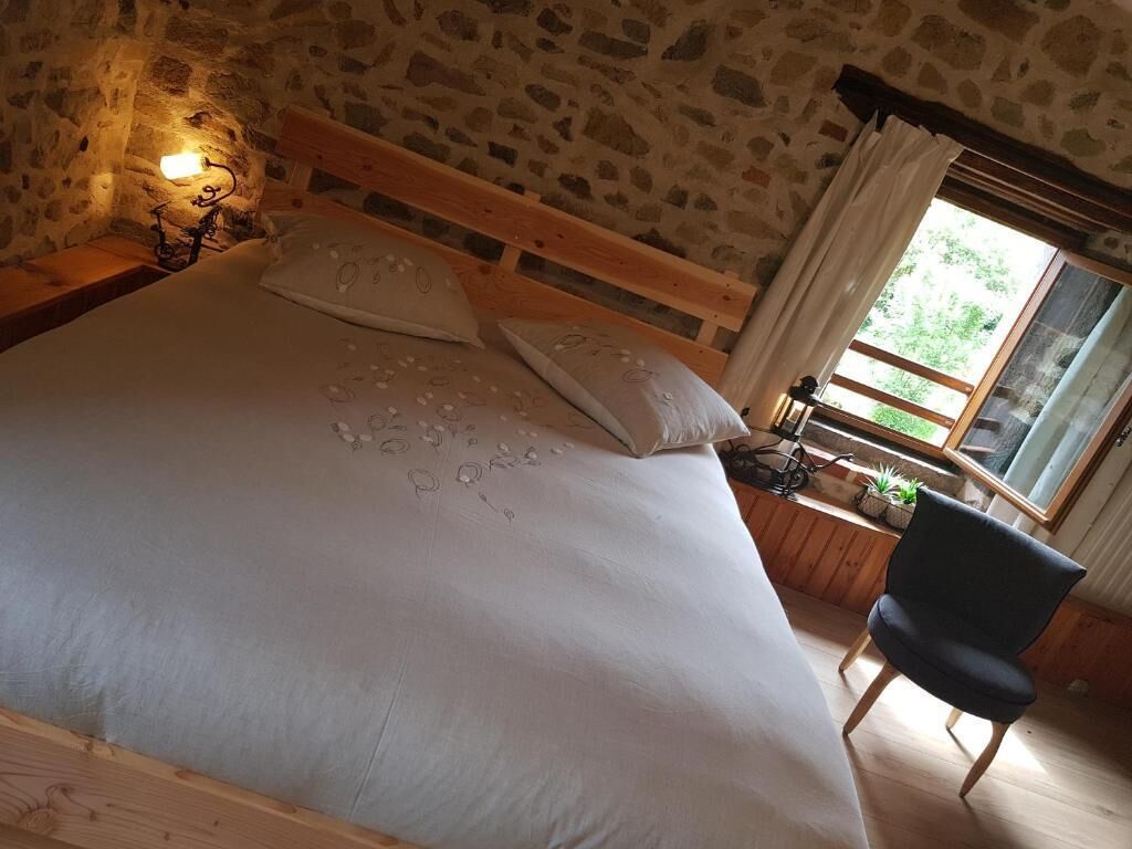 Un dormitorio con pared de piedra, estructura de cama de madera, mesita de noche con lámpara, una silla y ventana con cortinas.