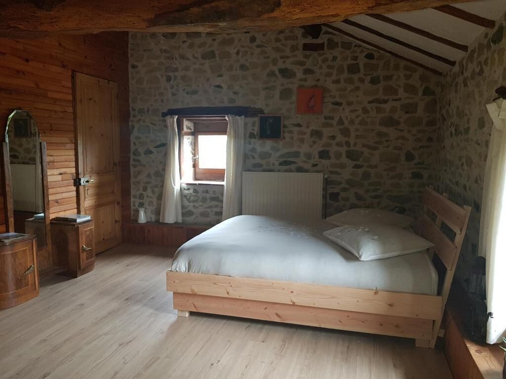Une pièce rustique avec un mur en pierre, un plancher en bois, un lit avec un cadre en bois, une fenêtre avec des rideaux et une porte en bois.