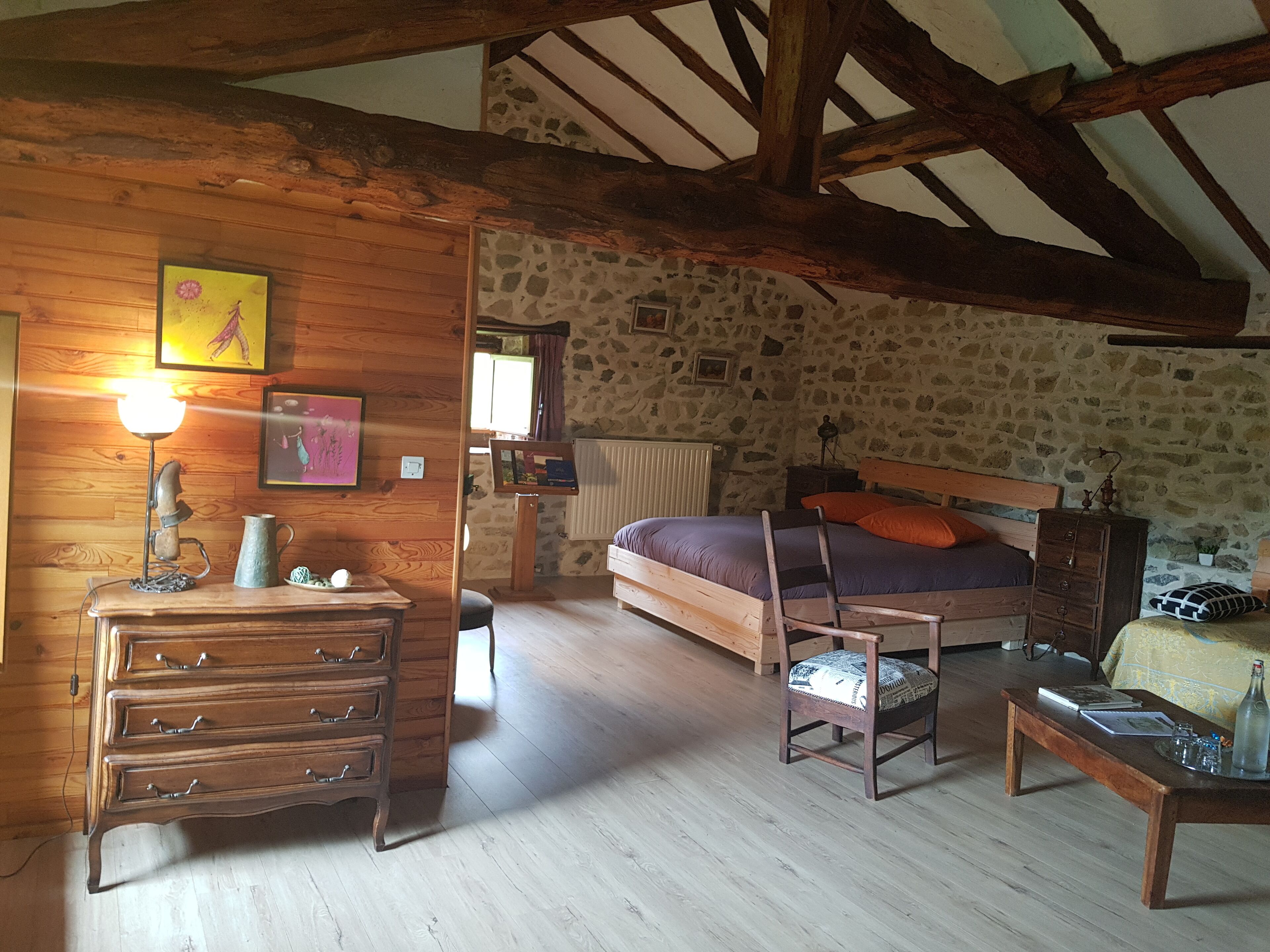 A rustic bedroom with a wooden dresser, a bed with a wooden headboard, a stone wall, and a wooden ceiling beam.