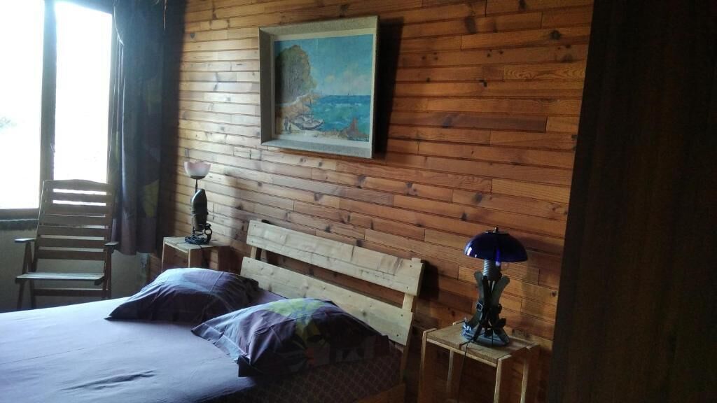 A room with a wooden wall, a bed with a headboard, a chair, a lamp, and a painting.