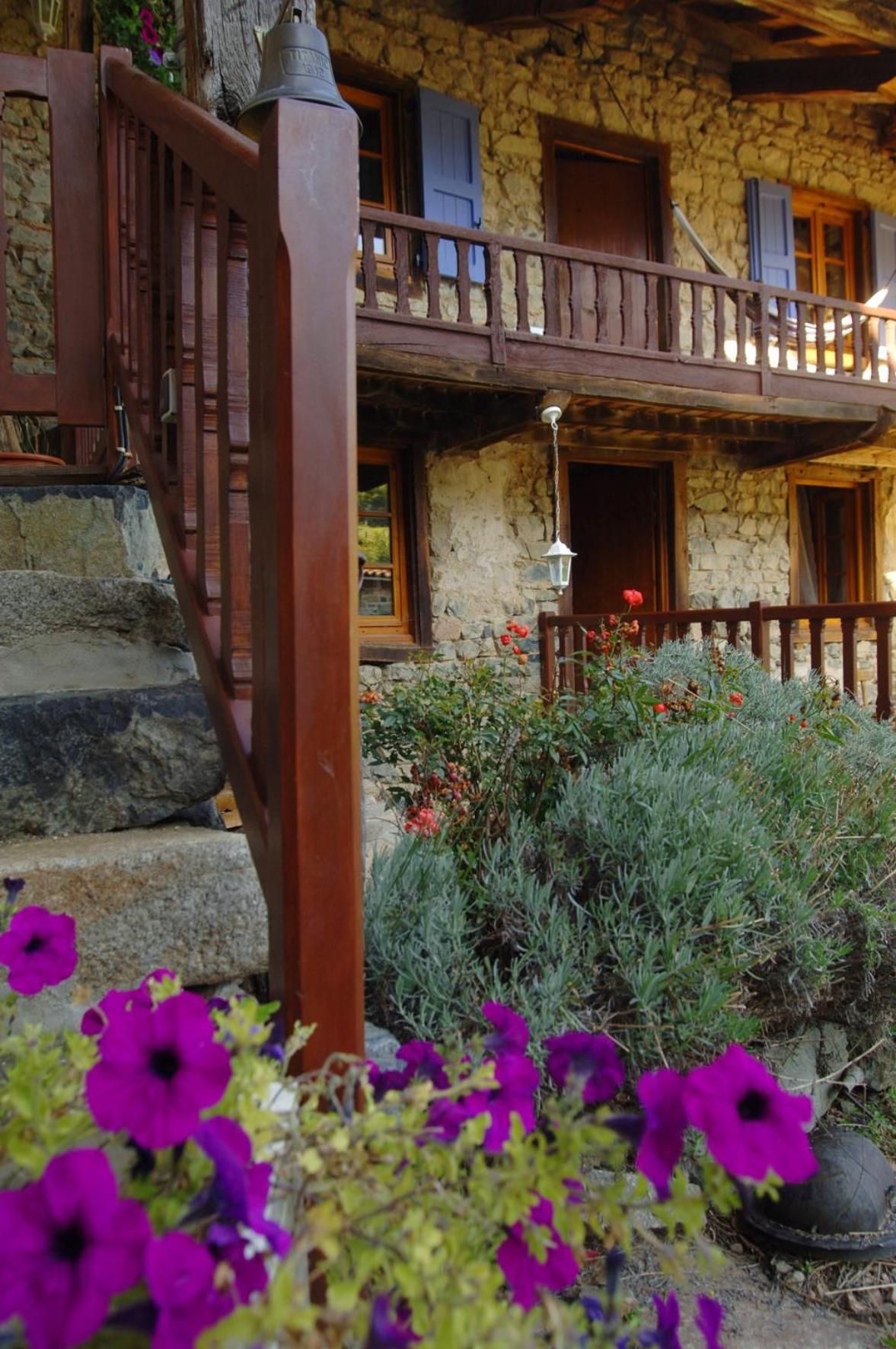 Un edificio de piedra con un balcón de madera, escalones que conducen a la entrada y un jardín con flores moradas.