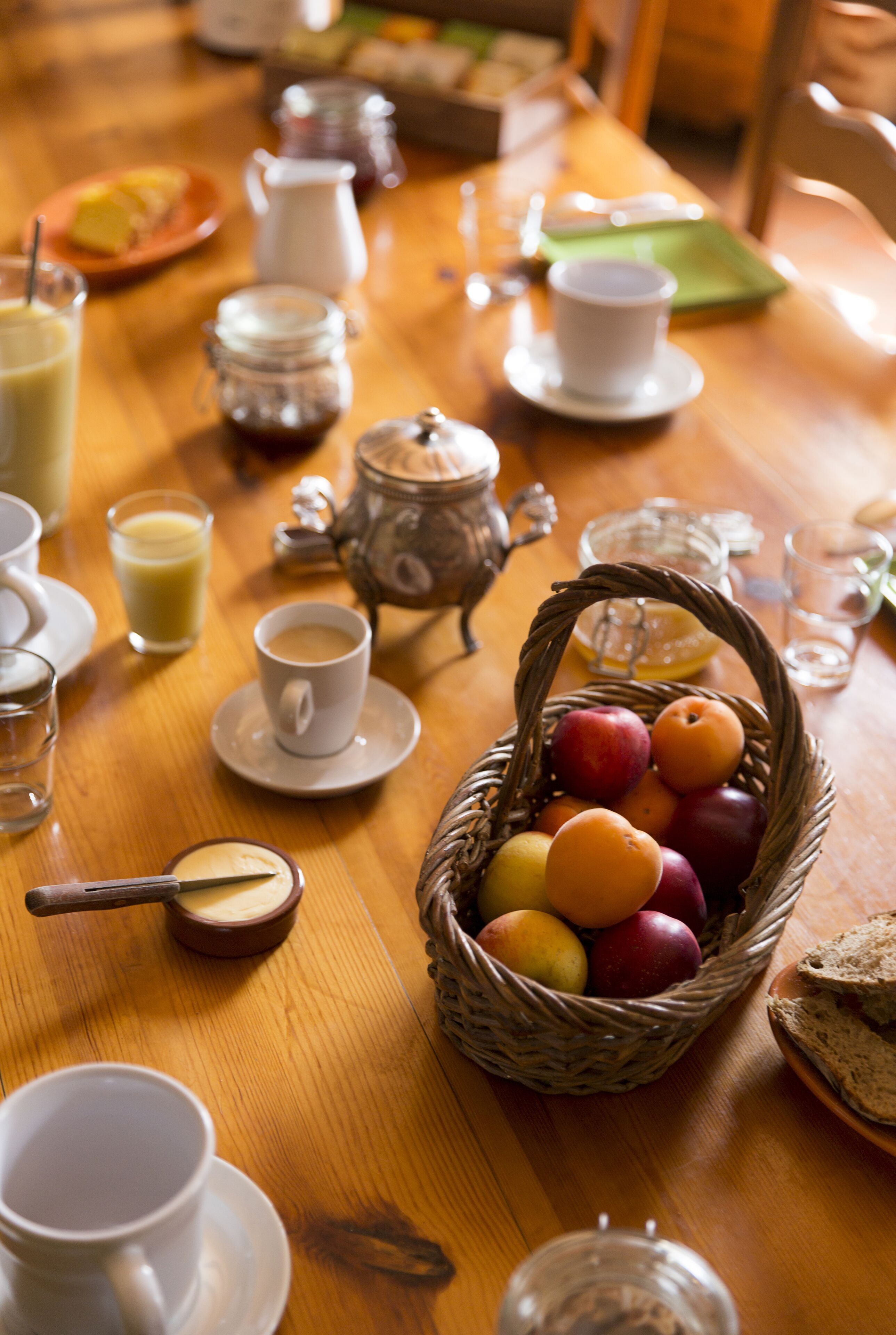 Una mesa de madera con alimentos para el desayuno, incluyendo una cesta de fruta, tazas de café y una tetera.