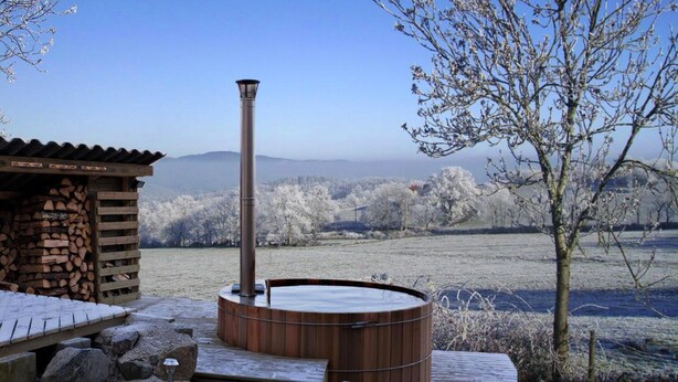 A wooden hot tub with a chimney, a stone wall, and a view of a frosty landscape.