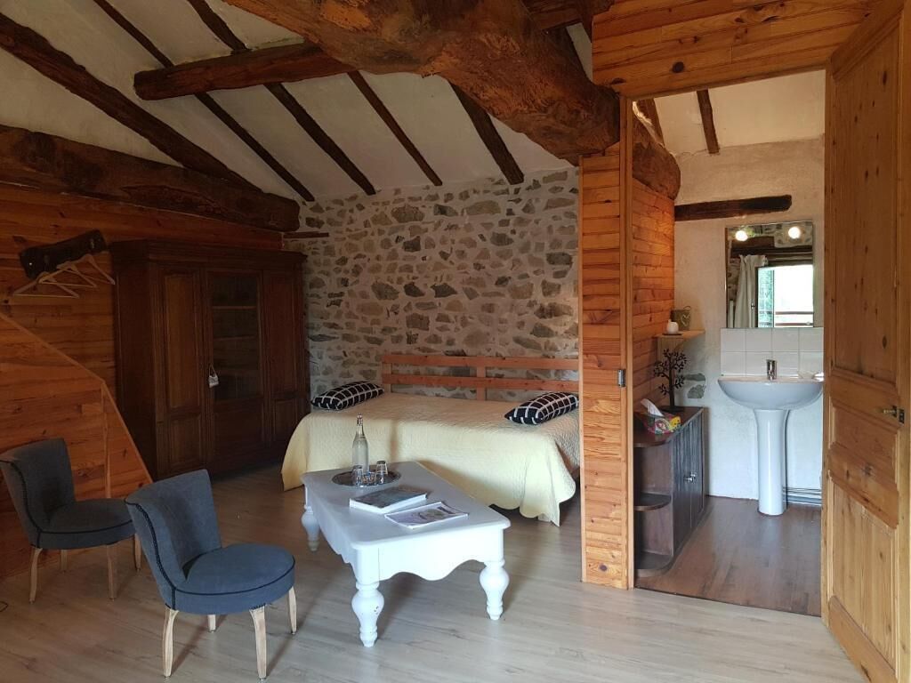 Una habitación acogedora con pared de piedra, vigas de madera, una cama, una mesita, una silla y un baño con lavamanos y espejo.