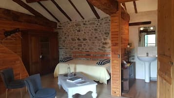 Una habitación acogedora con pared de piedra, vigas de madera, una cama, una mesita, una silla y un baño con lavamanos y espejo.