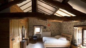 A rustic bedroom with a stone wall, wooden beams, a bed, a chair, and a small table.