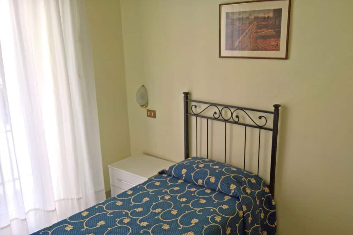 Une chambre à coucher avec un lit, une table de chevet, un tableau encadré au mur et une fenêtre avec des rideaux.