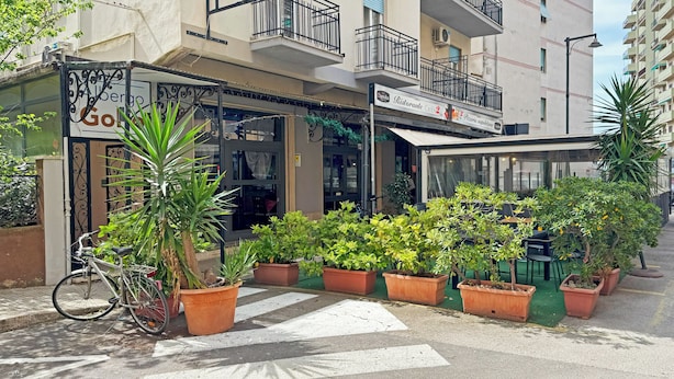 Un edificio con un letrero que dice "Alberto Goldoni" y "Pizzeria napoliana". Hay macetas con plantas frente al edificio.