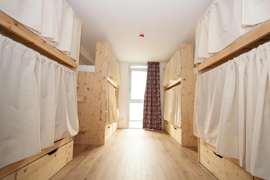 A narrow hallway with wooden bunk beds on either side, leading to a window with a colorful curtain.