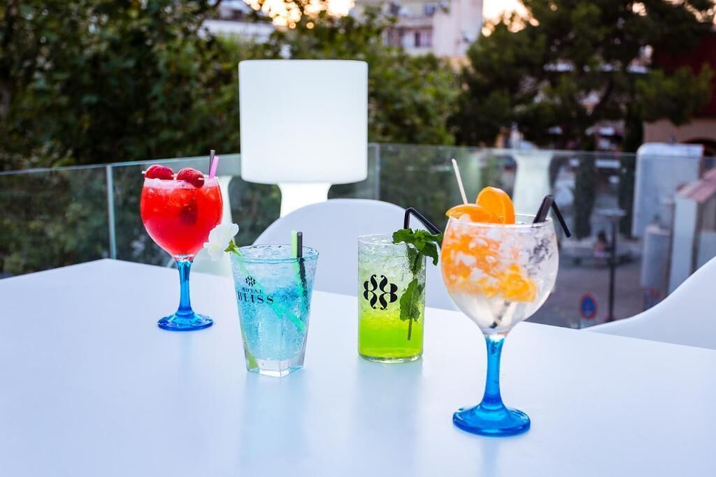A table with four colorful cocktails on a white table, set outdoors with a view of trees and buildings in the background.