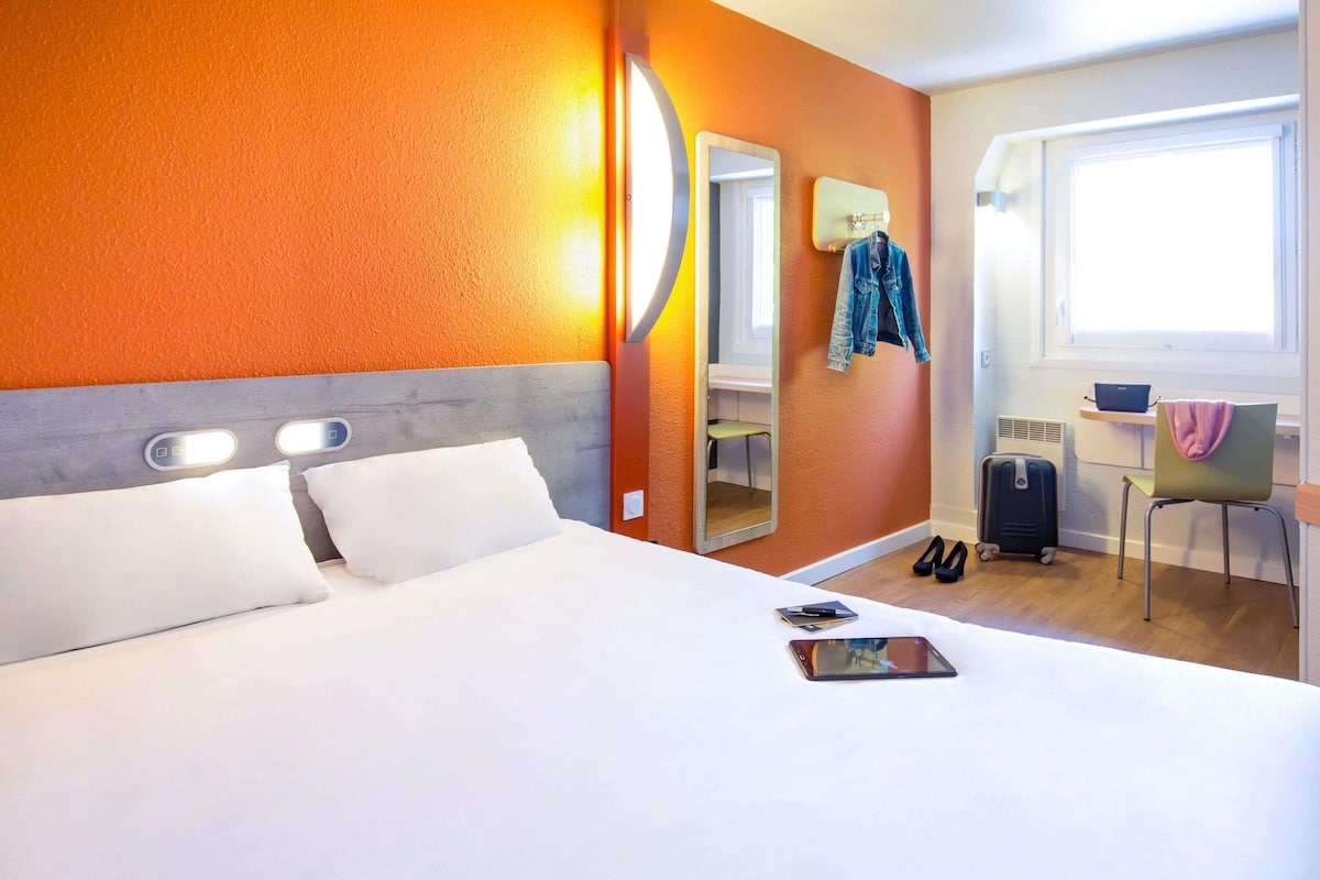 Una habitación de hotel con cama, escritorio, silla, maleta y espejo.