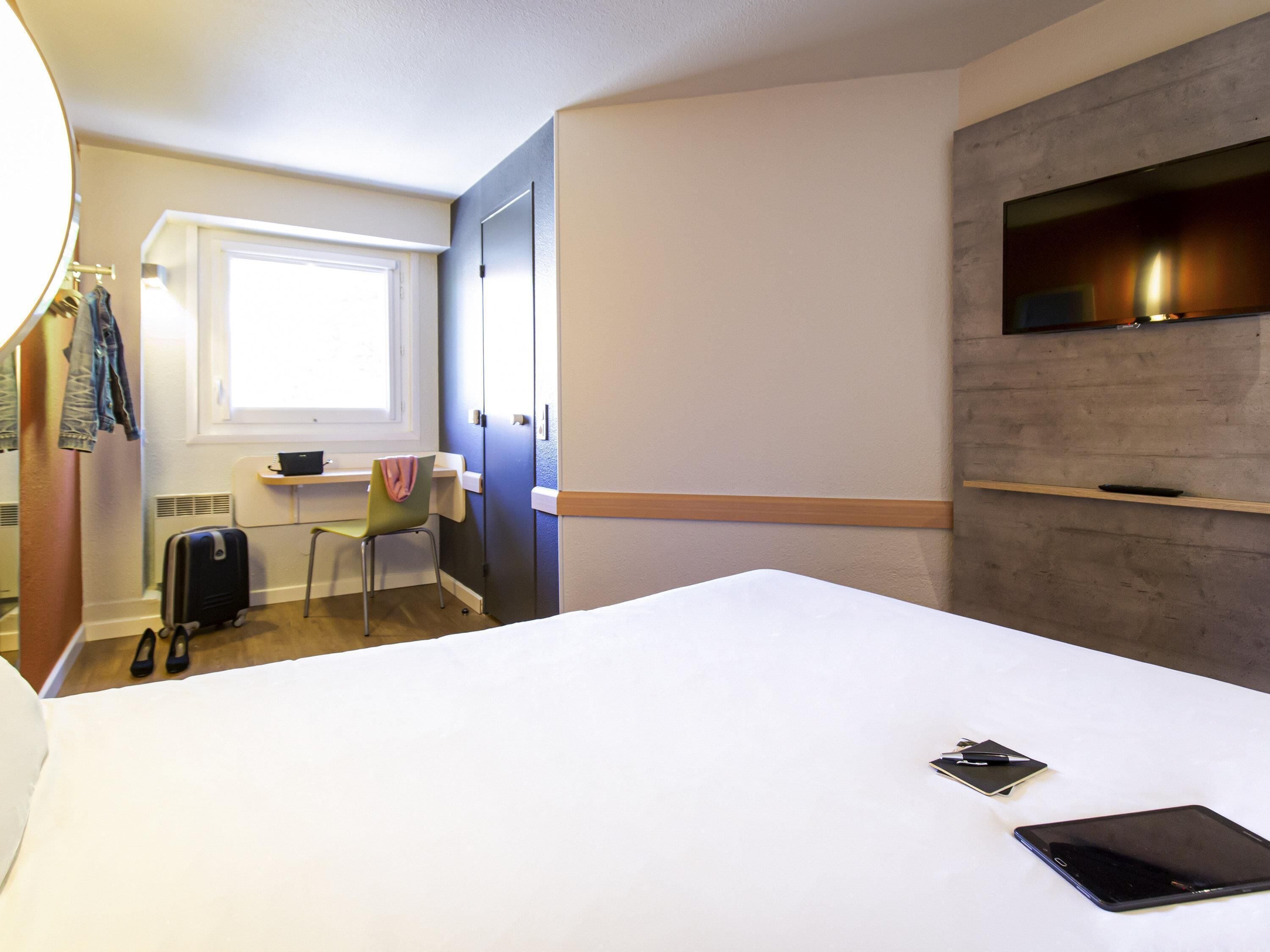 A hotel room with a bed, a desk, a chair, a suitcase, and a TV mounted on the wall.
