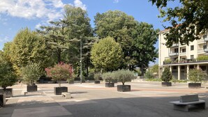 An outdoor urban area with a paved plaza, several potted trees, and a building with balconies in the background. - ibis budget Vienne Sud (Vienne)
