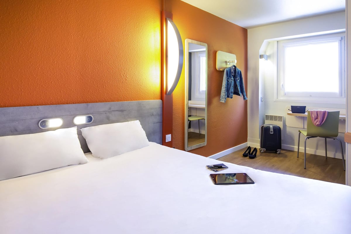 Una habitación de hotel con cama, escritorio, silla, maleta y espejo.