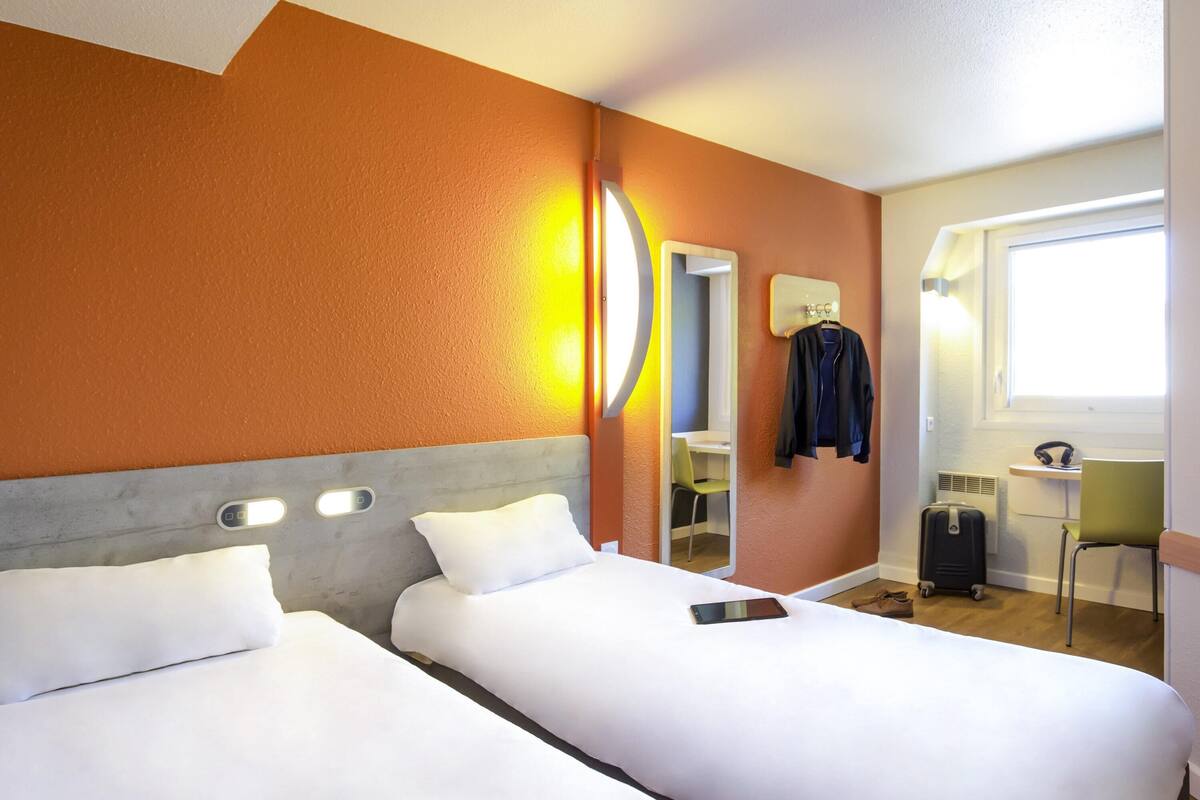 A hotel room with two beds, a desk, a chair, a suitcase, and a window.