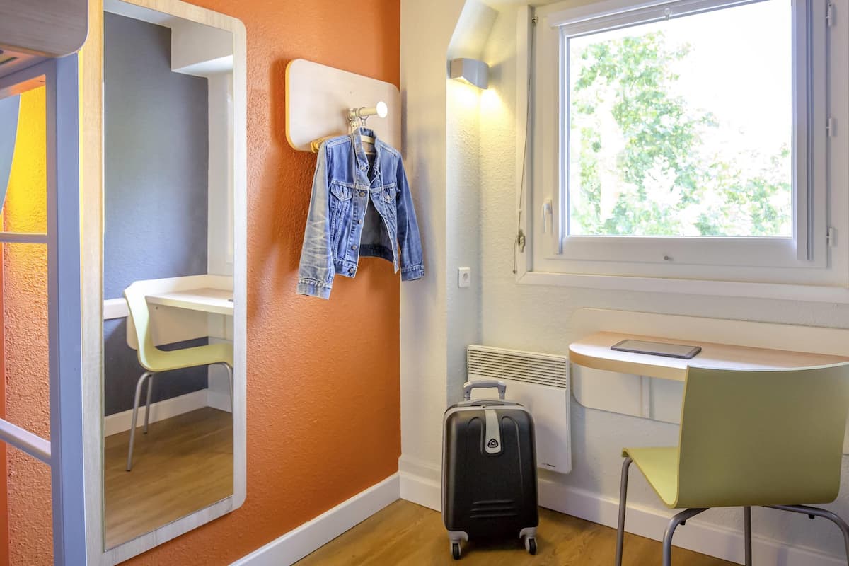 A hotel room with a mirror, a desk, a chair, a suitcase, and a window with a view of trees.