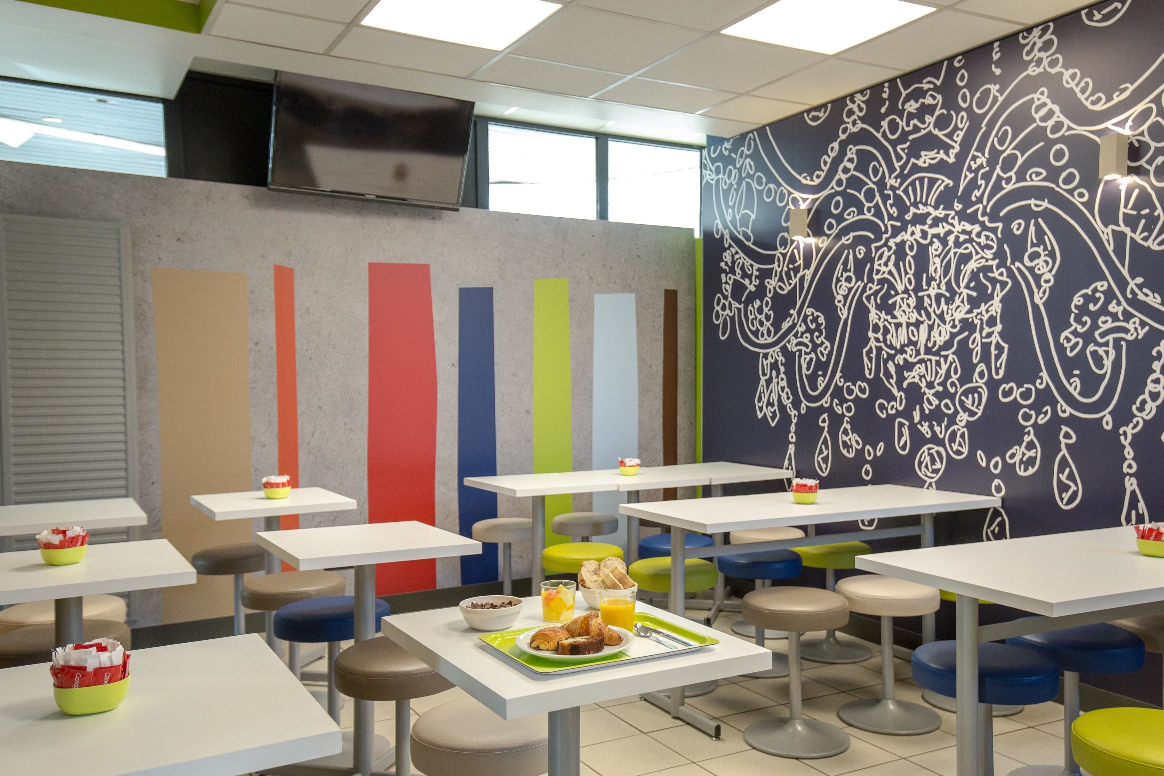 Una cafetería con mesas blancas y sillas coloridas, un muro con estampado y un televisor montado en la pared.
