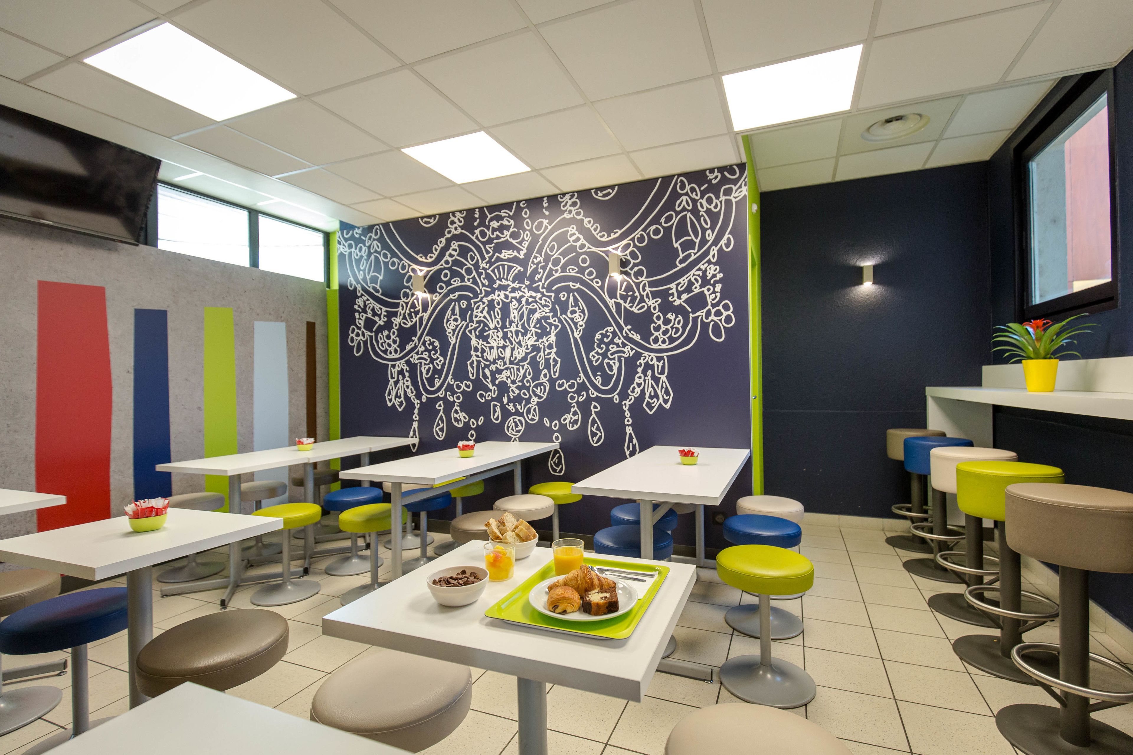 A brightly lit cafeteria with multiple tables and stools, featuring a colorful wall mural and a potted plant.
