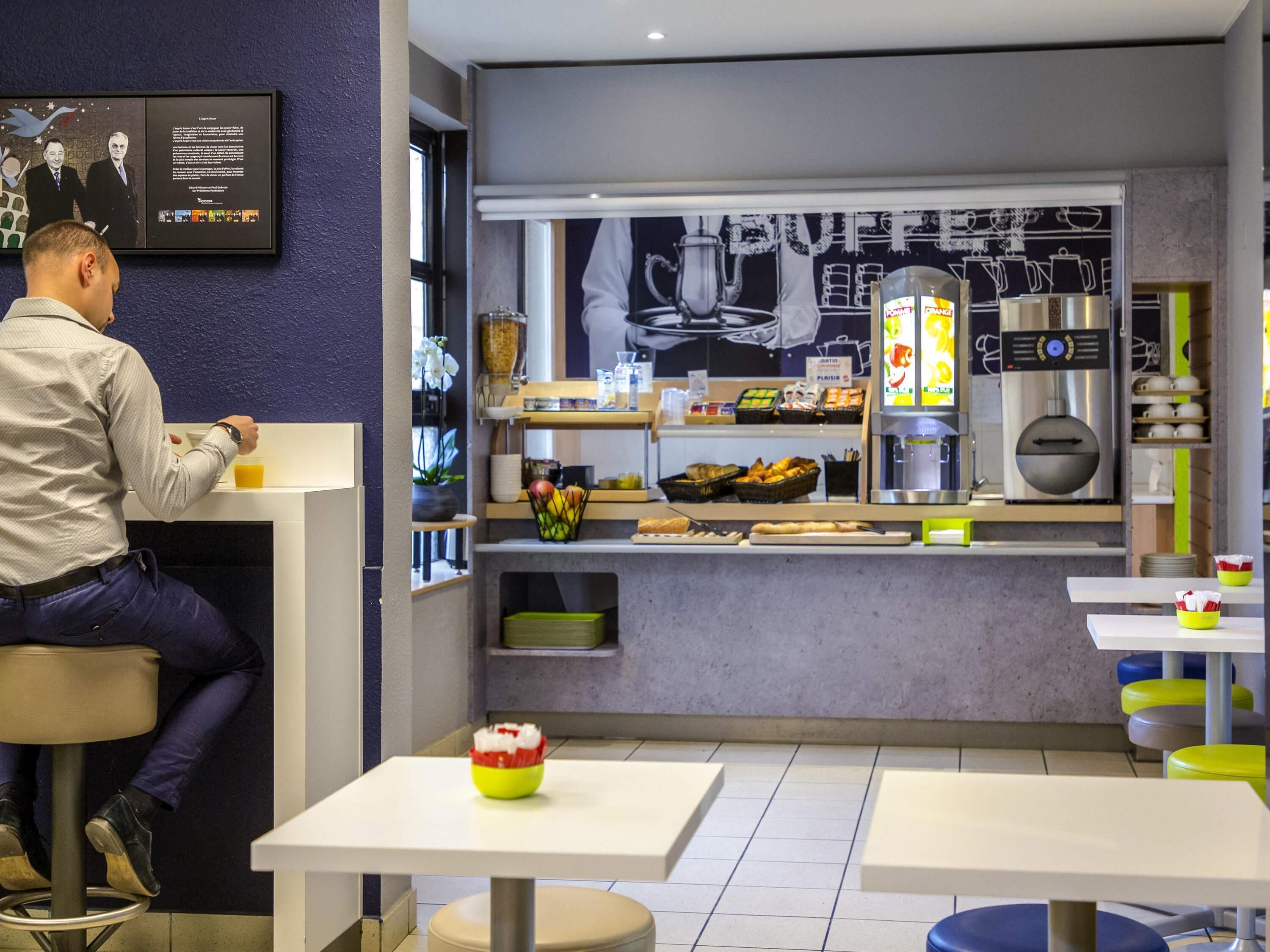 A modern cafe with a counter displaying pastries and coffee machines. There are tables and chairs for seating.