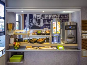A breakfast bar with a coffee machine, fruit, pastries, and a chalkboard menu. - ibis budget Vienne Sud (Vienne)