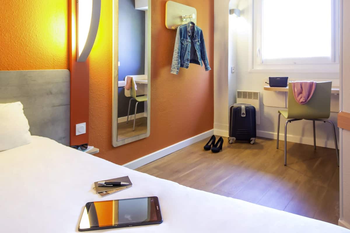 A hotel room with a bed, a desk, a chair, a suitcase, and a mirror on the wall.