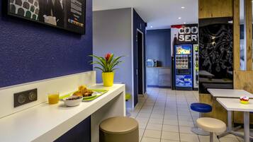 Un moderno vestíbulo de hotel con mostrador de recepción, área de descanso, máquina expendedora y un televisor empotrado que muestra un paisaje urbano.