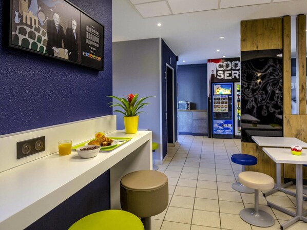 Un moderno vestíbulo de hotel con mostrador de recepción, área de descanso, máquina expendedora y un televisor empotrado que muestra un paisaje urbano.