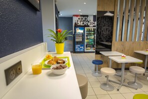 A breakfast area with a counter, a plant, a beverage, and a vending machine in the background. - ibis budget Vienne Sud (Vienne)