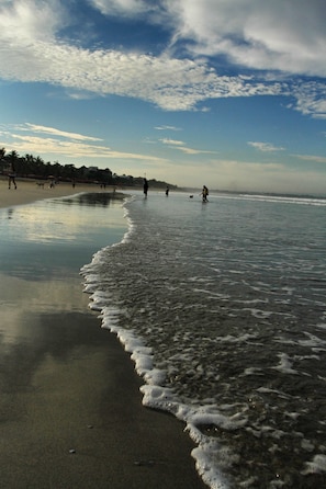 A beach with people walking along the shore, waves gently lapping at the sand, and a clear sky above.