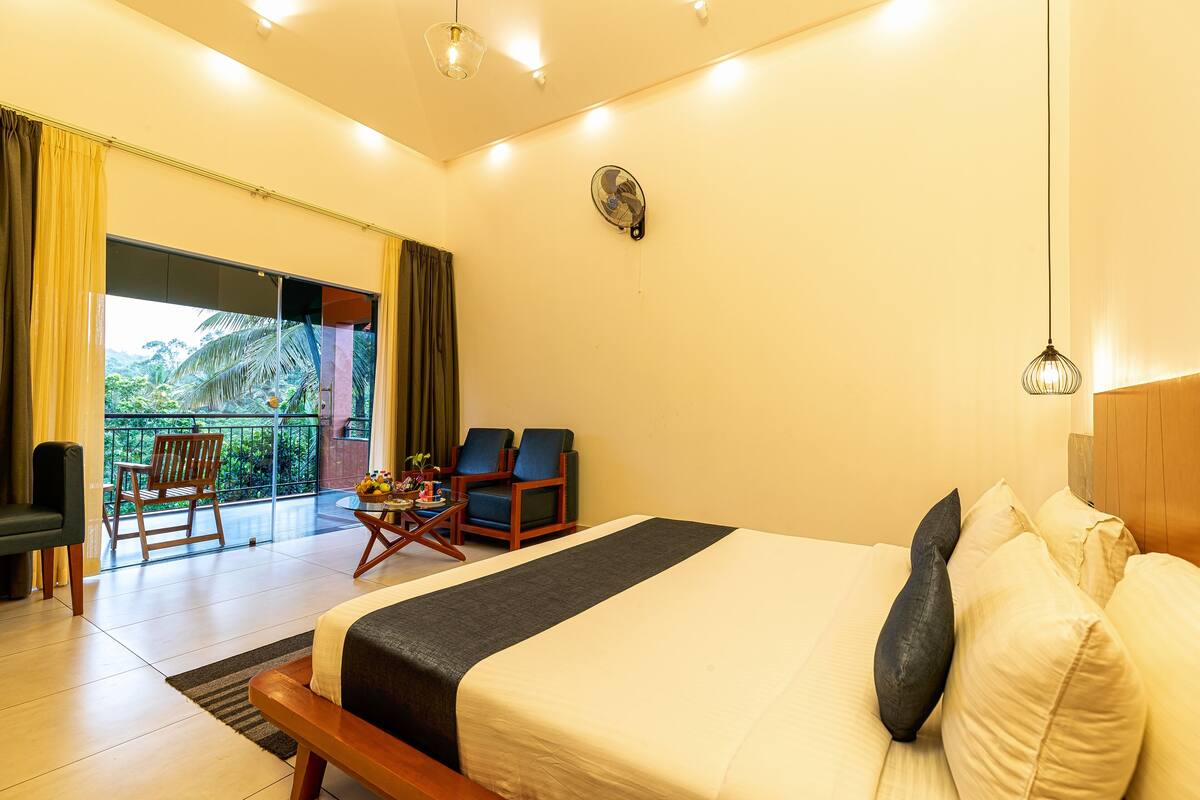 A hotel room with a large bed, a small table, two chairs, and a balcony with a view of greenery.