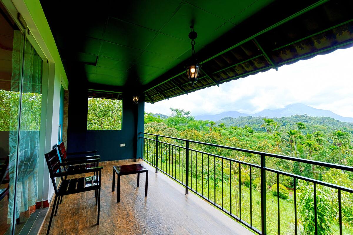 A balcony with a wooden floor, a table, and chairs, overlooking a lush green landscape with mountains in the distance.